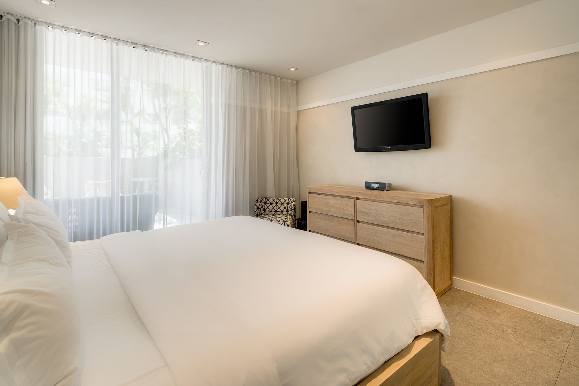 A wide view of a hotel room with a large bed, a dresser with a TV on top, and a window with sheer curtains.