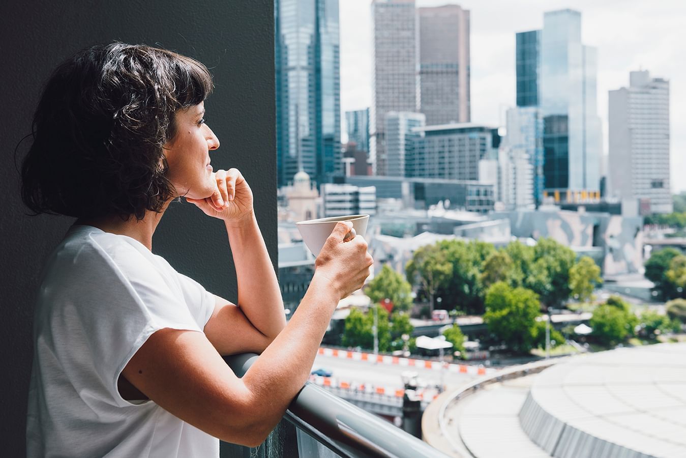 Lady overlooking the city view while drinking coffee from a balcony at Quay West Suites Melbourne