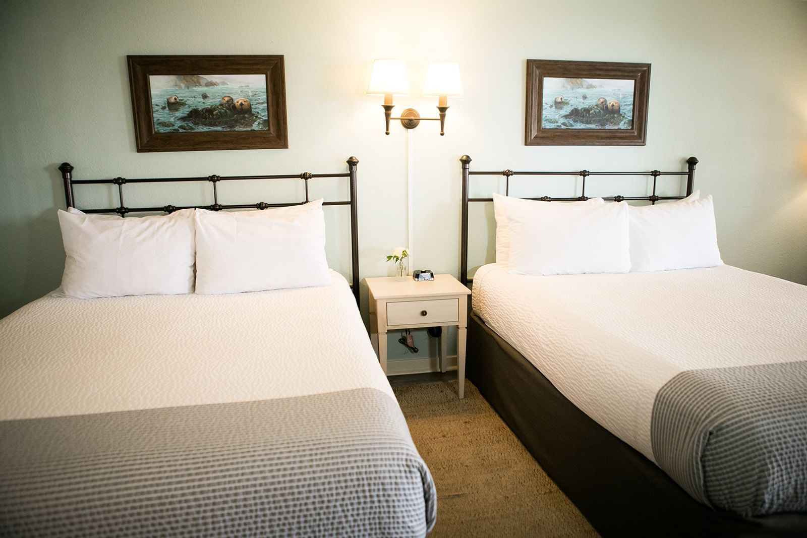 A hotel room with two queen beds, a nightstand, and artwork on the walls.