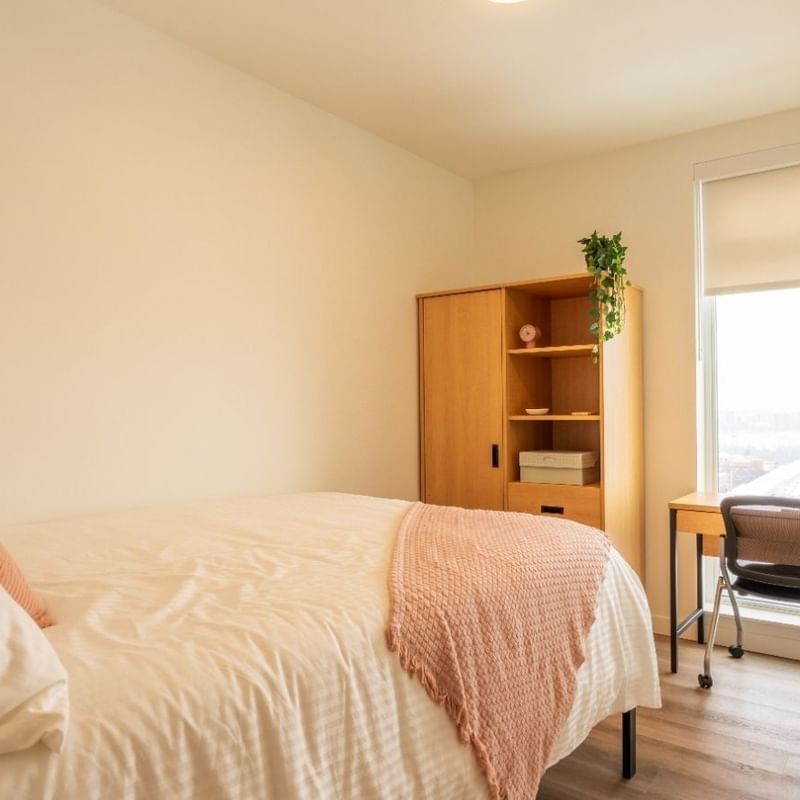 A light-filled bedroom with a bed, floor to ceiling windows, a desk, a desk chair, a dresser and a wardrobe. 