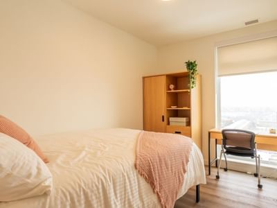 A light-filled bedroom with a bed, floor to ceiling windows, a desk, a desk chair, a dresser and a wardrobe. 
