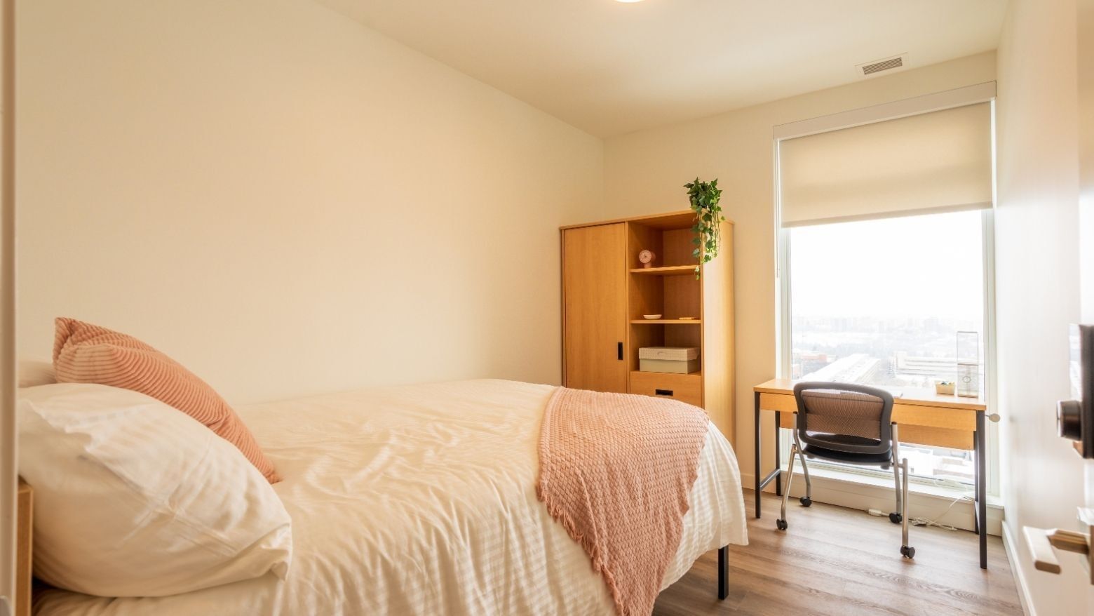 A light-filled bedroom with a bed, floor to ceiling windows, a desk, a desk chair, a dresser and a wardrobe. 