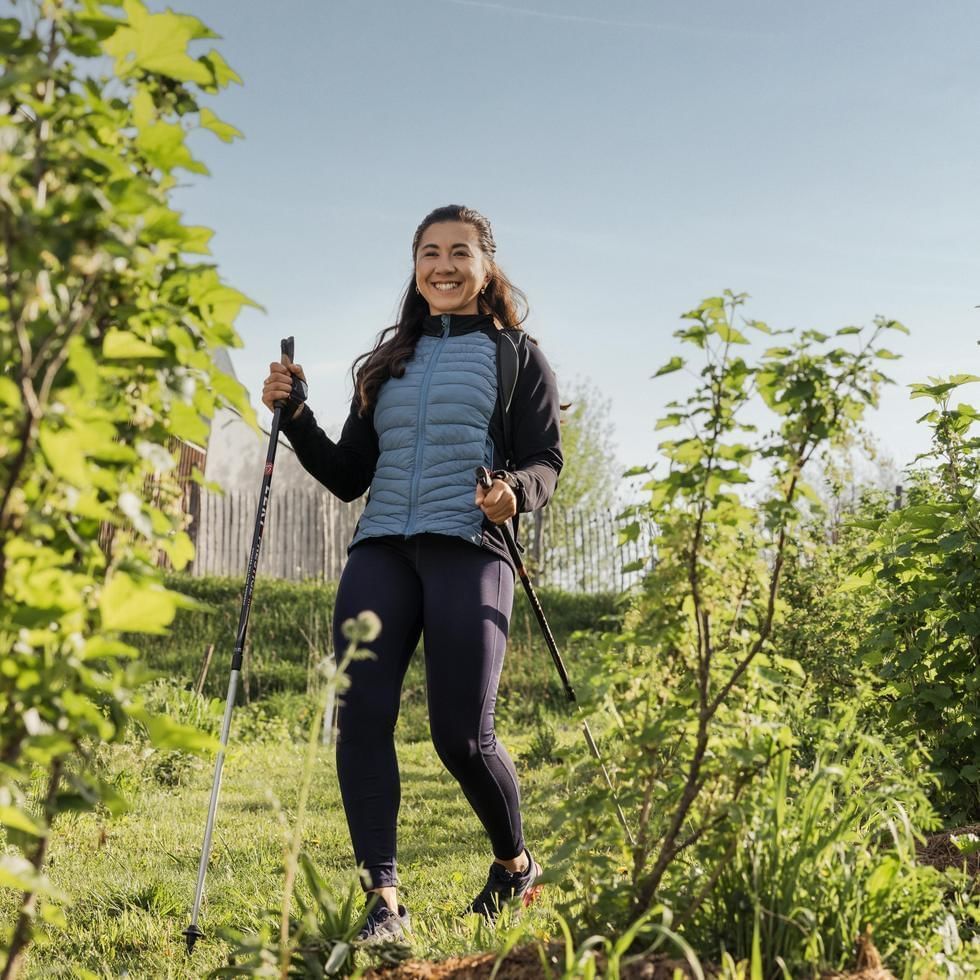 Woman smiling with trekking poles in a grassy area with green plants and trees in the background.