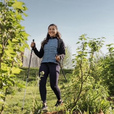 Woman smiling with trekking poles in a grassy area with green plants and trees in the background.