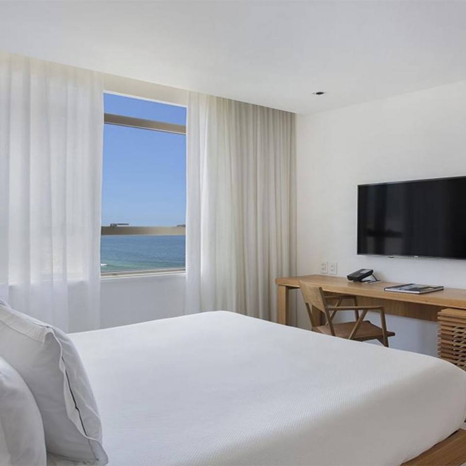 Suíte com vista para o mar do Janeiro Hotel Leblon RJ decorada com cama, bancada com cadeira e TV
