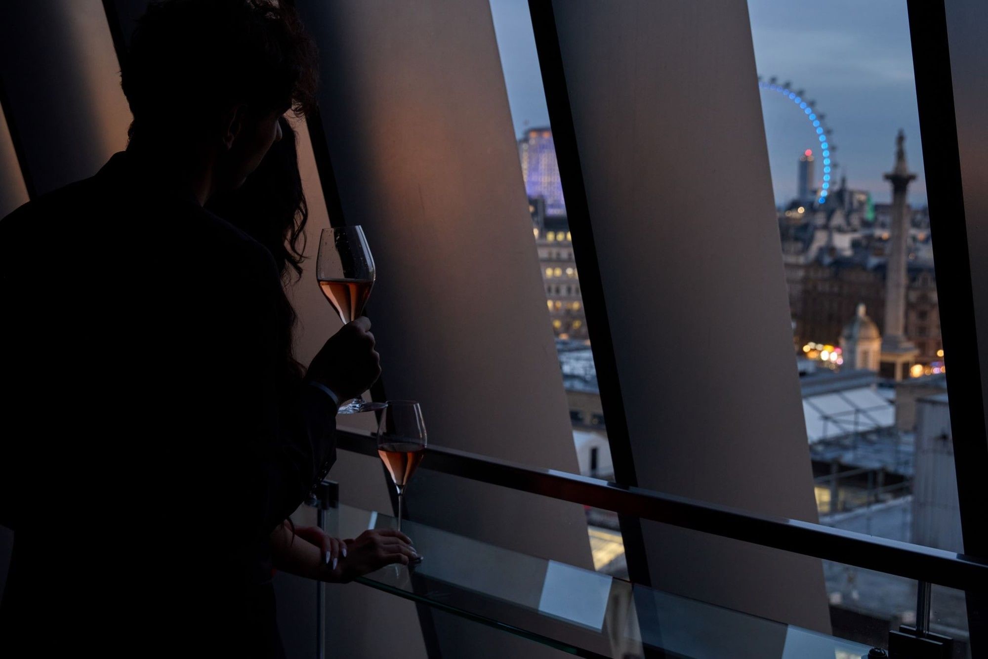 Silhouetted couple holding glasses of rosé wine while looking out at the London Eye from Flannigans at The Londoner Hotel