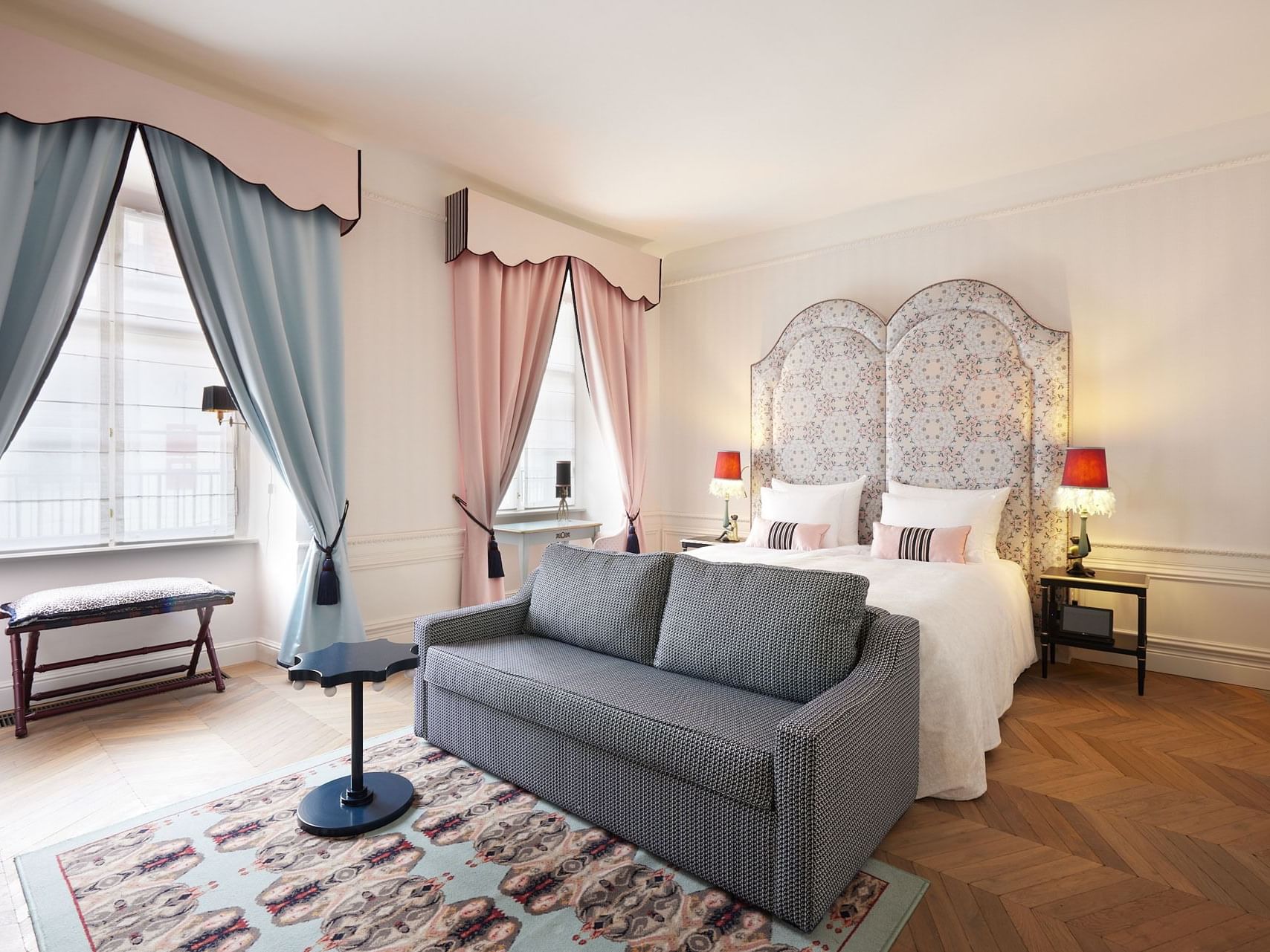 A bedroom with a large bed, ornate headboard, patterned rug, gray couch, and two windows with drapes.