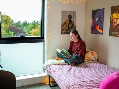 Student reading on bed in bright room with large window overlooking green trees and decorative posters on wall