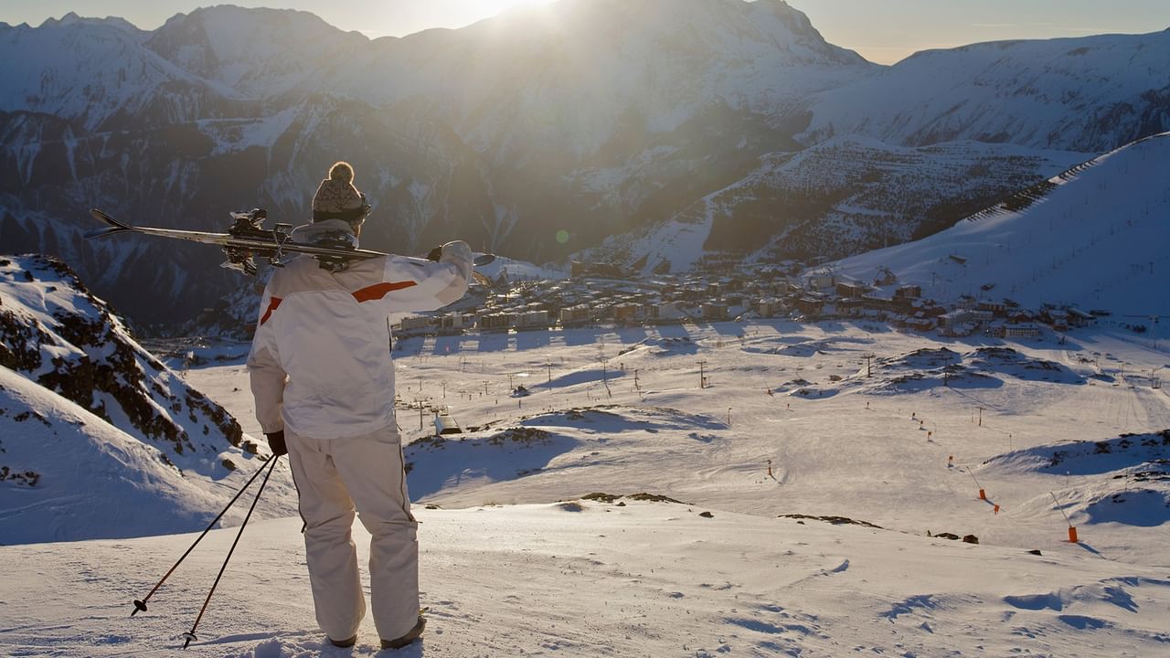 Skier carrying skis and poles while overlooking a snow-covered mountain valley and ski resort