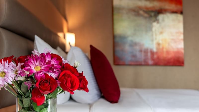 Hotel room with a bed, pillows, and a vase of flowers next to a wall painting.