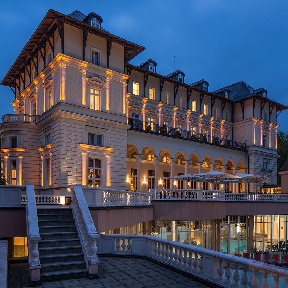 Noční pohled na osvětlený hotel s bazénem a terasou s deštníky.