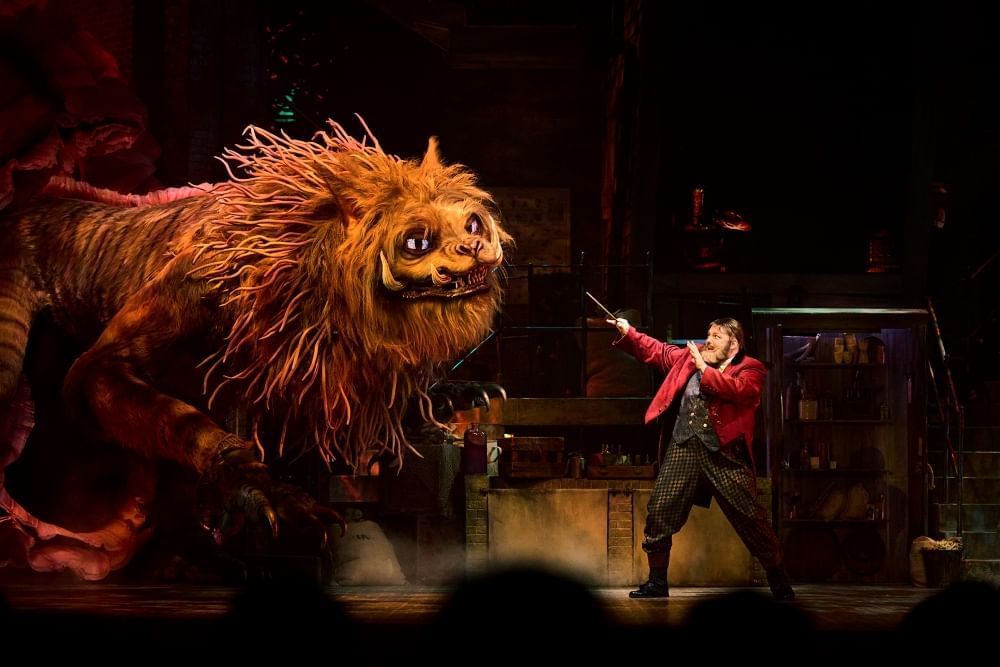 A large mysterious beast with a man towers over a man in a red jacket, pointing a wand at the creature.