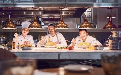 Three chefs posing at the Opera kitchen counter with large plates of noodles and rice in Park Hyatt Saigon