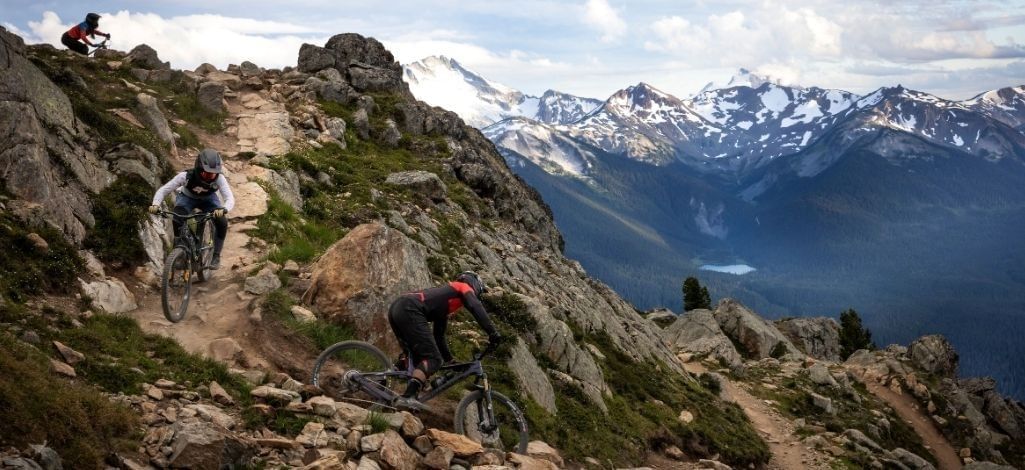 Mountain bikers descend a rocky alpine trail with snow-capped peaks in the background.
