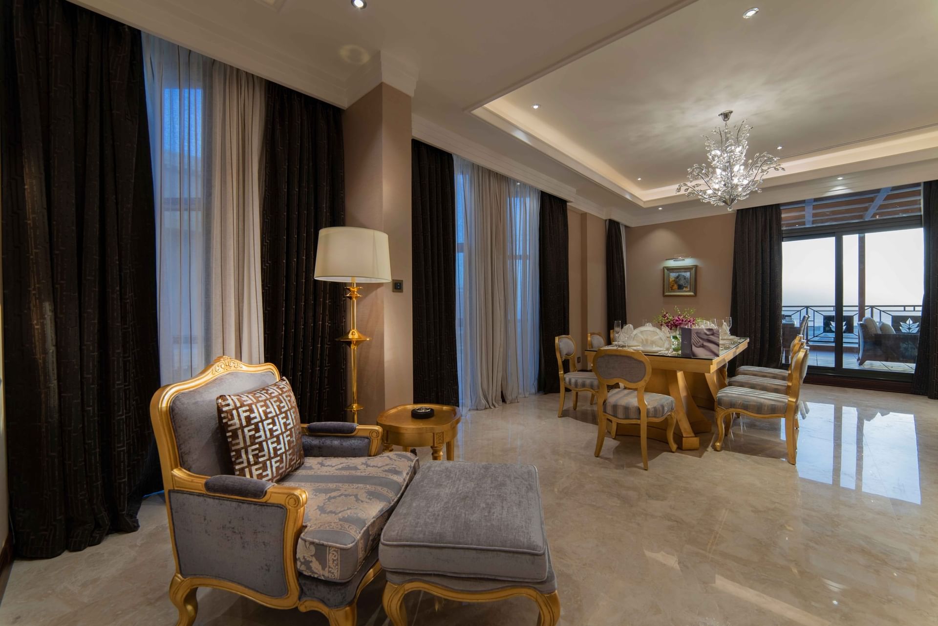 Gilded armchair & ottoman by a formal dining table under a massive crystal chandelier in Penthouse Villa at Warwick Al Khobar
