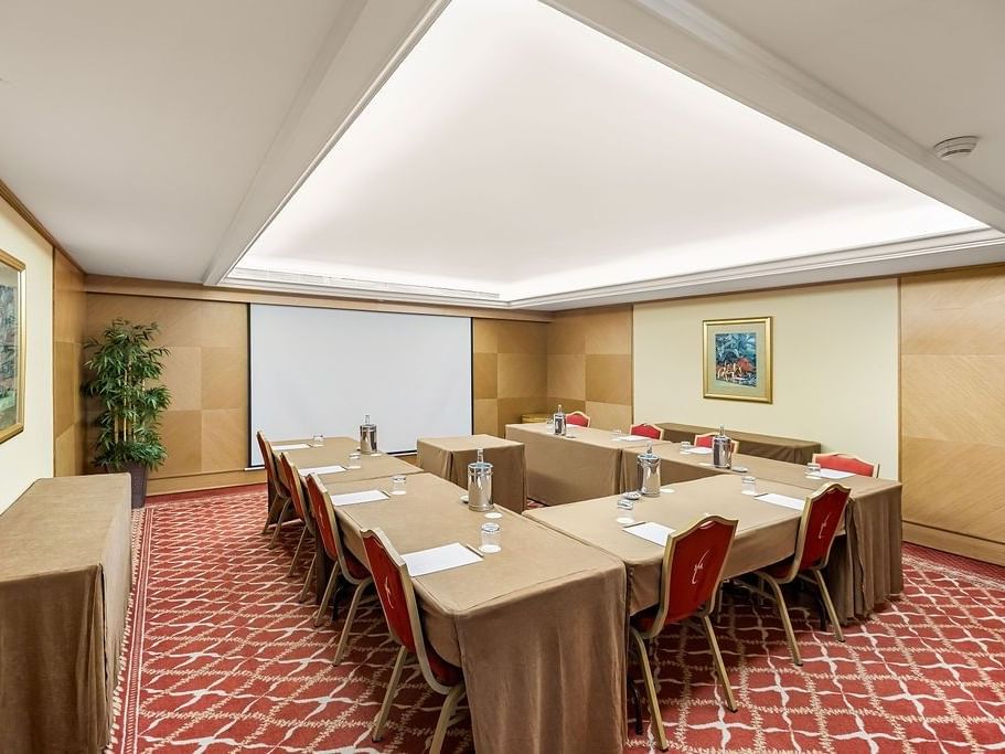 Sala de reuniones con mesas cubiertas, sillas rojas, pantalla y dos cuadros en paredes laterales.