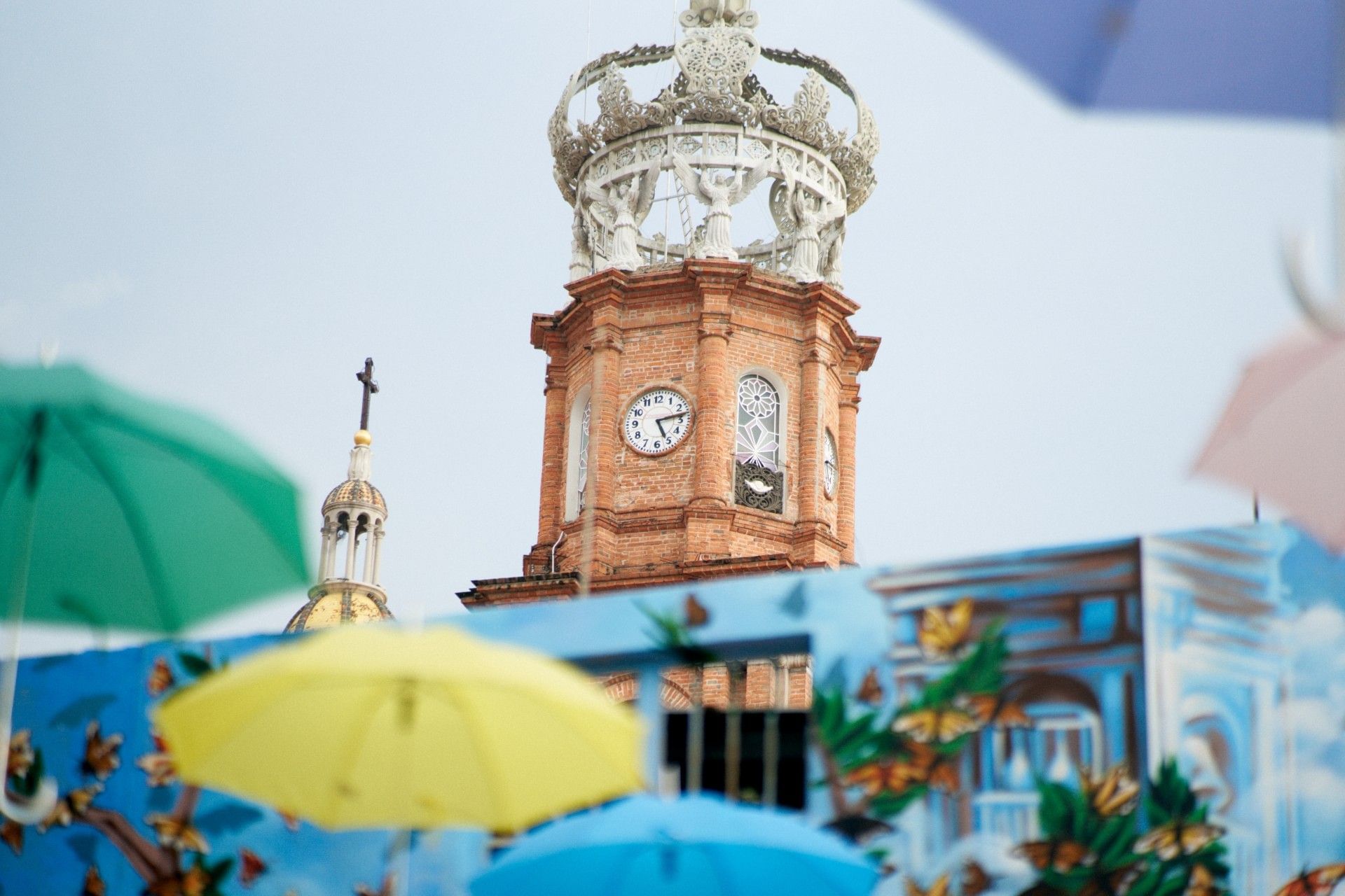 View of the Parish of Our Lady of Guadalupe tower in downtown Puerto Vallarta during whale season, with colorful umbrellas and urban art in the foreground.