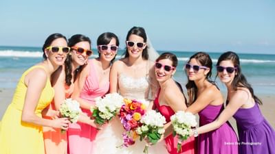 bride and bridesmaids on beach with sunglasses holding flower bo