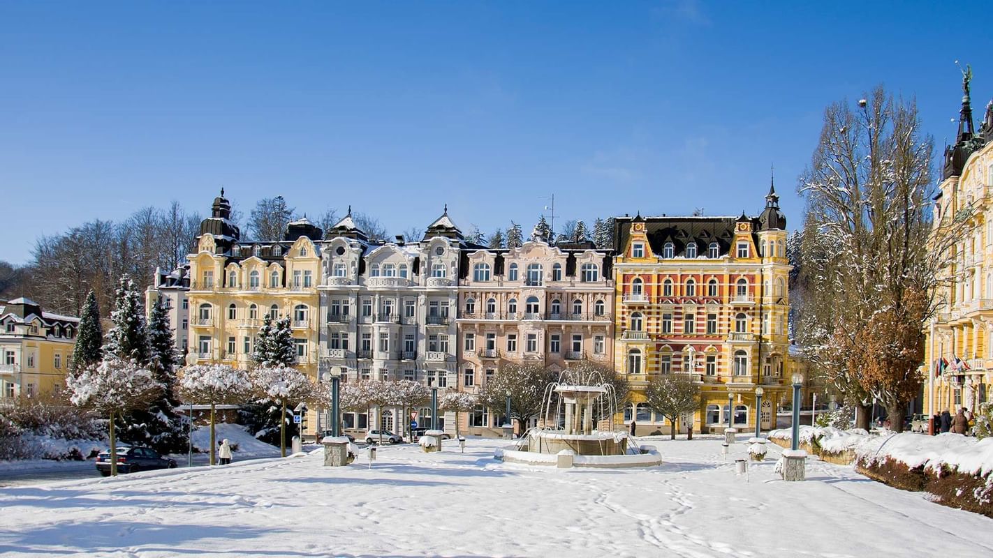 Schneebedeckter Platz mit Brunnen und bunten Gebäuden, umgeben von schneebedeckten Bäumen.