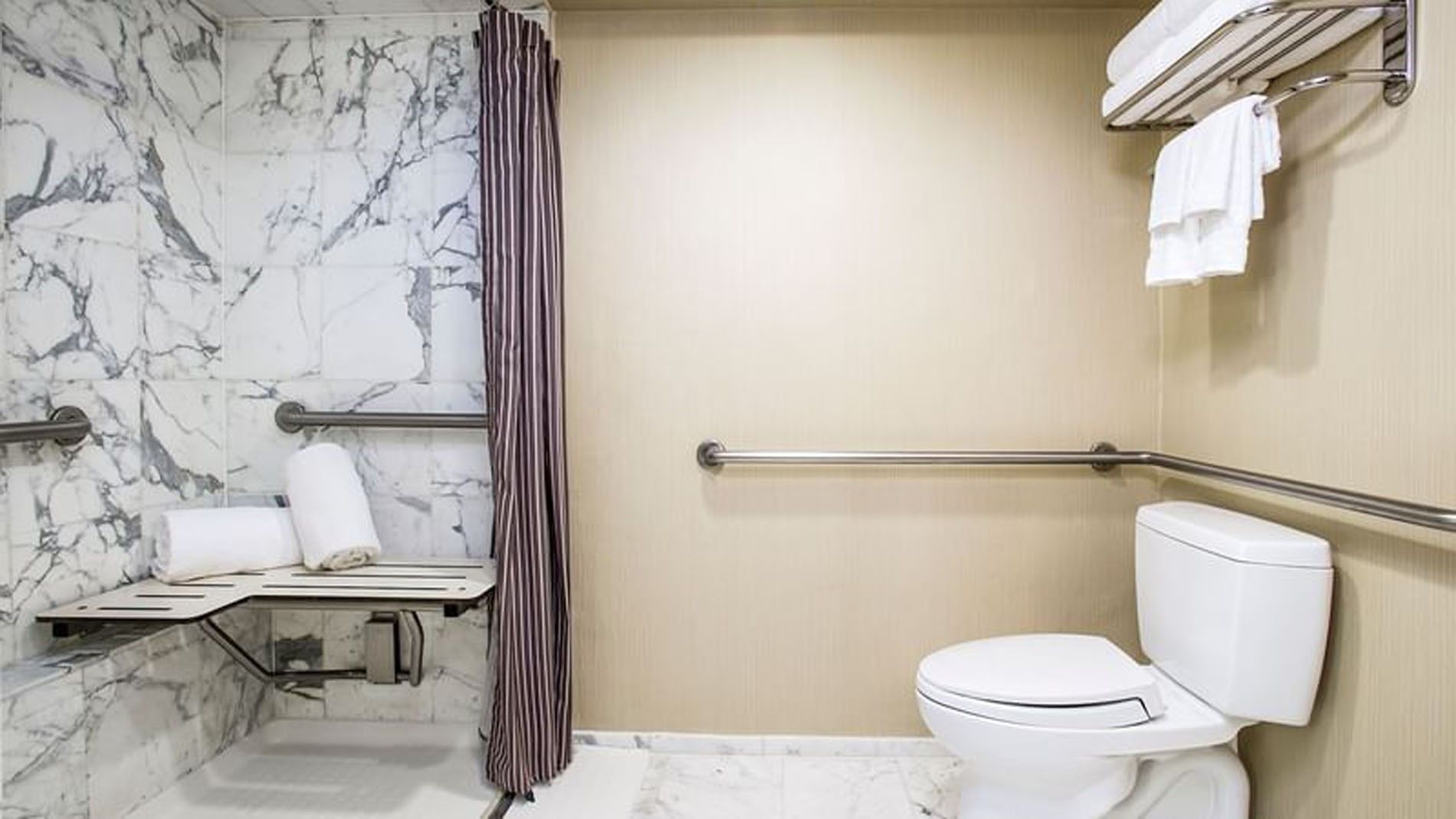 A bathroom with marble walls, a toilet, and a shower with a folding seat and grab bars.