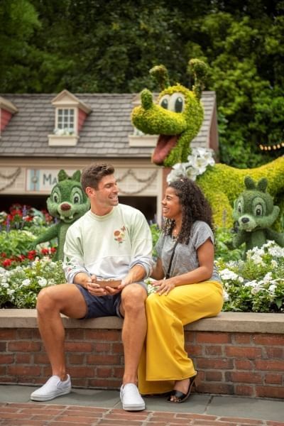 A man and woman sit on a brick ledge in front of topiary figures of Pluto, Chip, and Dale. 