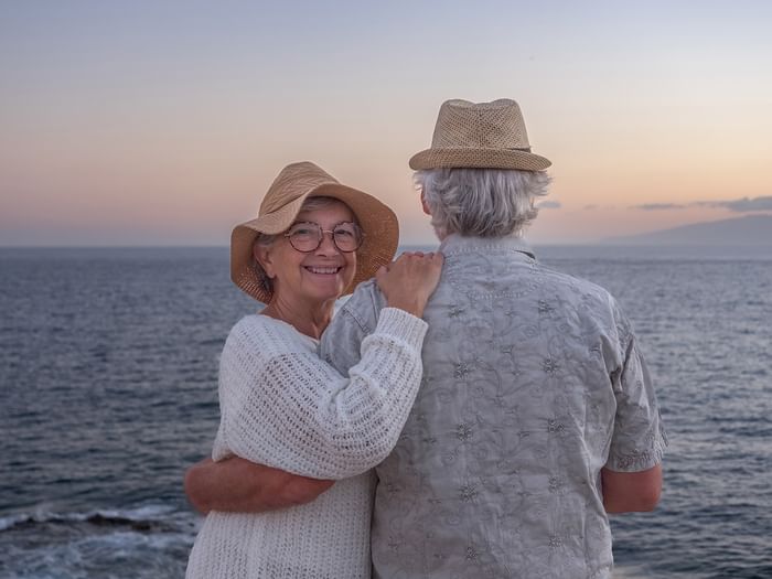 Elderly couple standing by the ocean at sunset, smiling and embracing each other.