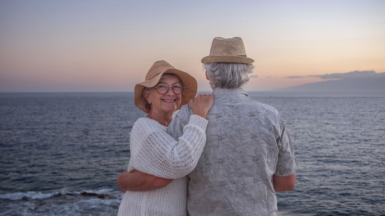 Elderly couple standing by the ocean at sunset, smiling and embracing each other.
