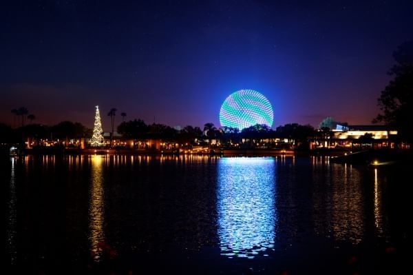 EPCOT's Spaceship Earth glowing brightly at night beside a lit Christmas tree across a lake.