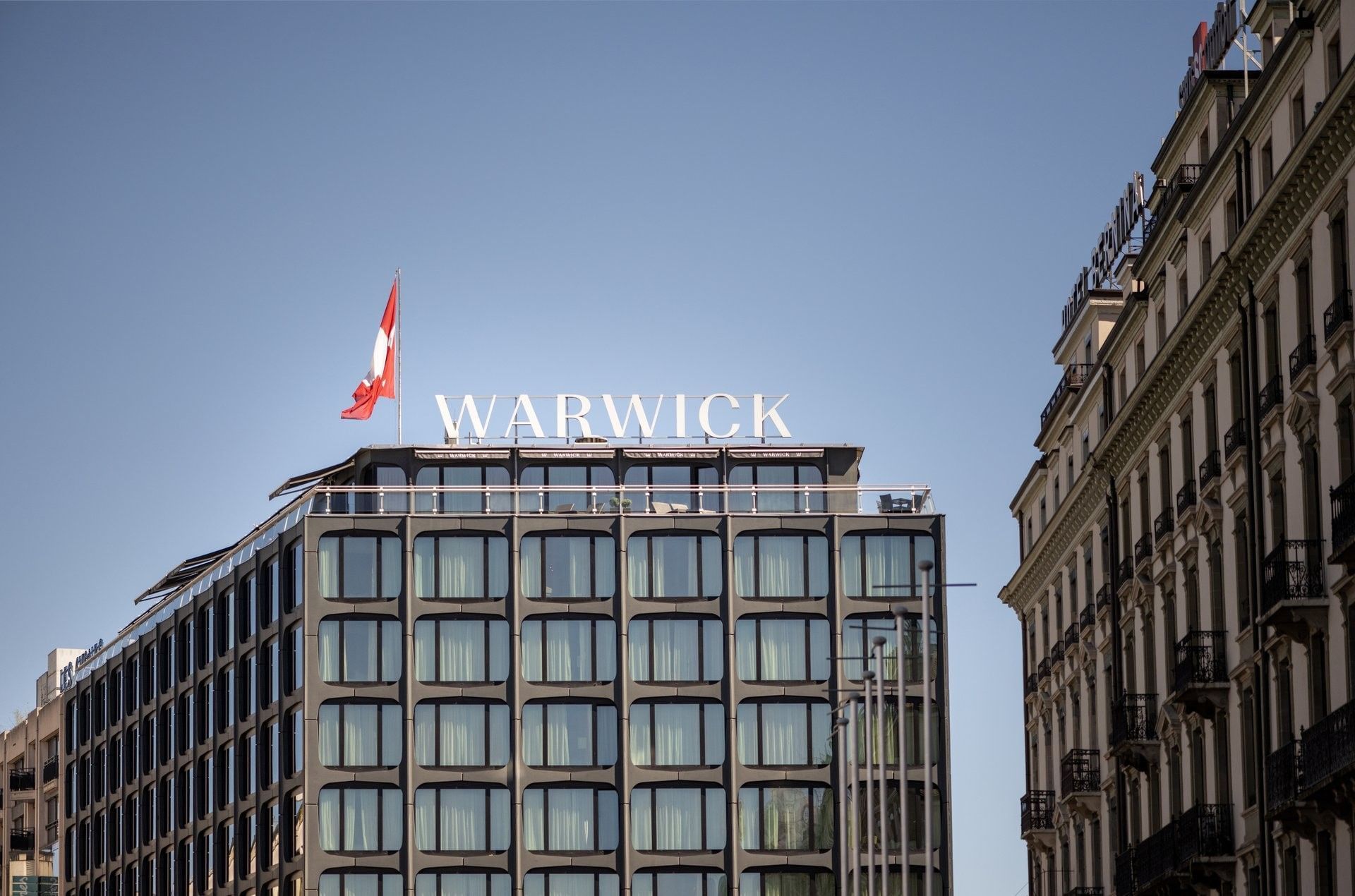 Exterior View of Warwick Geneva hotel with flag and rooftop signage under clear sky
