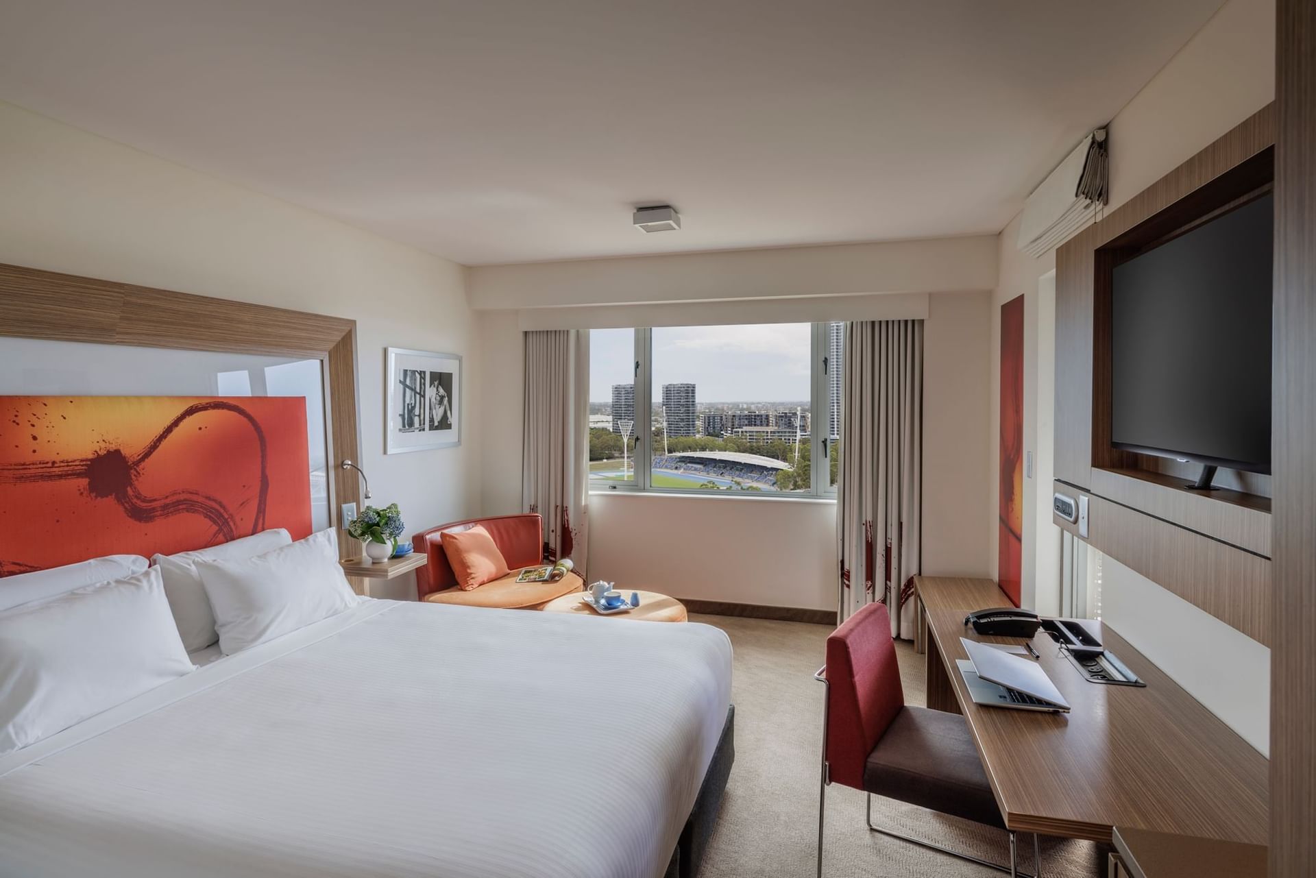 Superior Balcony room with a bed, desk with laptop, chair, TV, & large window with city view at Novotel Sydney Olympic Park