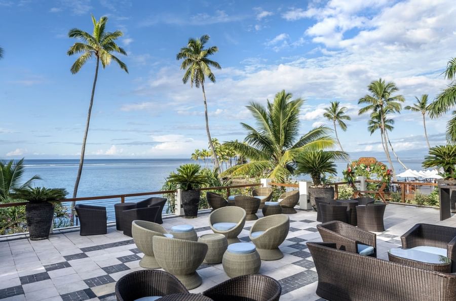 Round lounge chairs by a dark wicker sofa under palm trees in Sunset Terrace Bar at Warwick Fiji Resort and Spa
