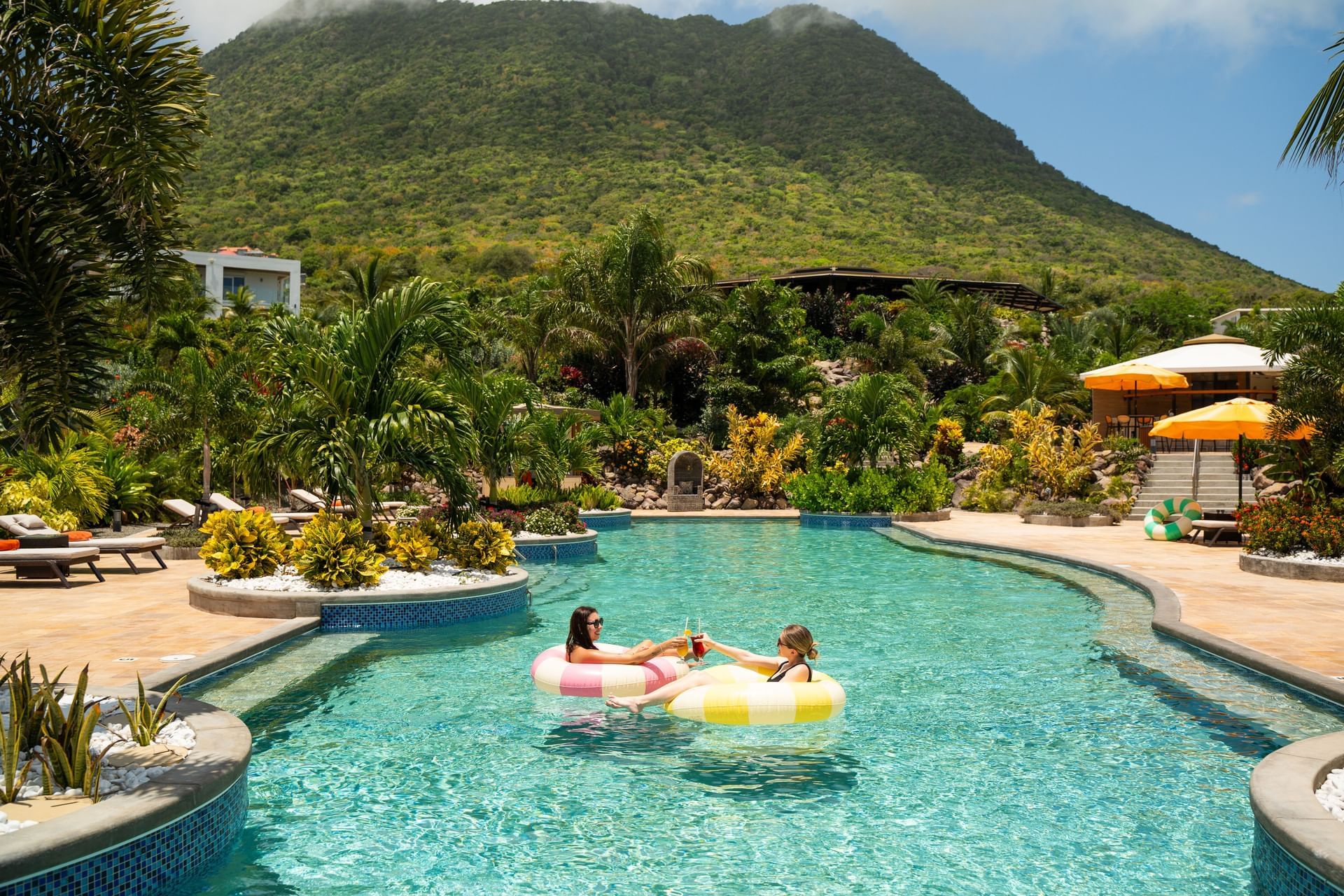 Twee vrouwen ontspannen in zwembanden in een zwembad, omringd door planten en een groene berg bij Golden Rock Resort
