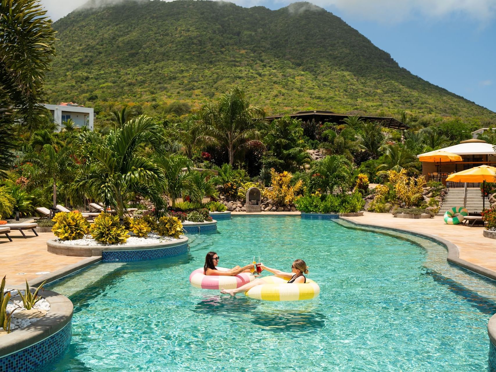 Two young women relax in inner tubes on a pool, surrounded by tropical plants and a lush mountain at Golden Rock Resort