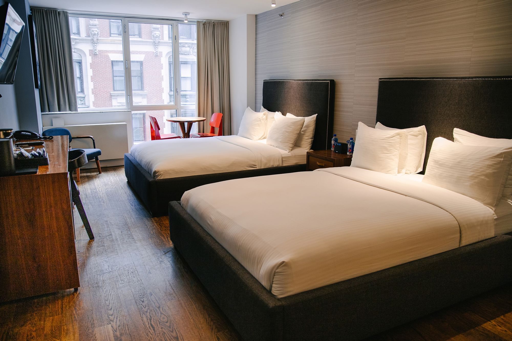 A hotel room with two beds, a desk, and a window with a city view.