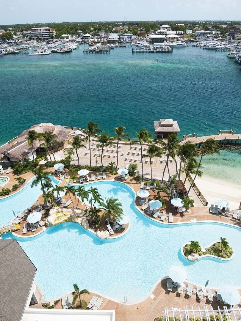 View of Warwick Paradise Island Bahamas, with a large pool by white umbrellas under palm trees surrounding a tropical harbor