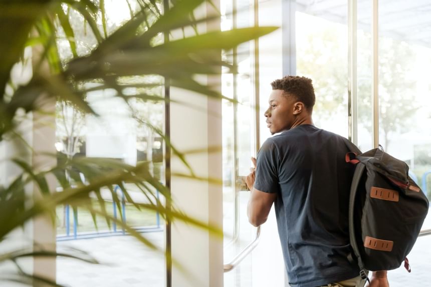 Man with a backpack walking by the glass entrance doors beside lush indoor greenery while traveling at Hotel Saint Clair