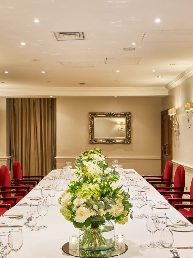 Elegant banquet room with long table, red chairs, floral centerpieces, and white walls.