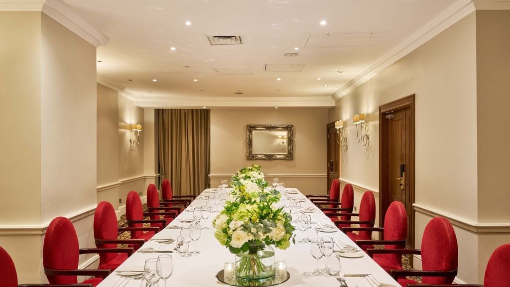 Elegant banquet room with long table, red chairs, floral centerpieces, and white walls.