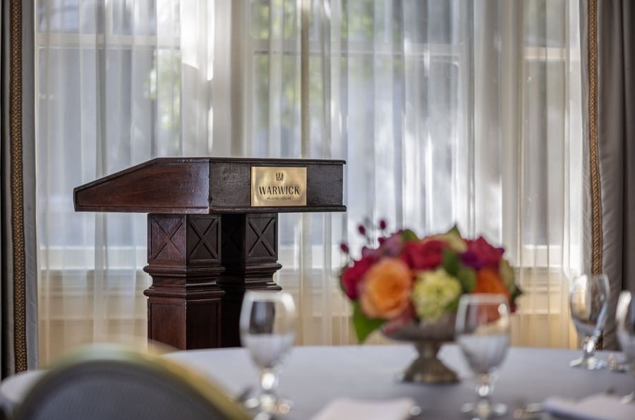 Wood podium by flowers under sheer white curtains in a sunlit conference in Oak Lawn/Terrace at Warwick Melrose Dallas