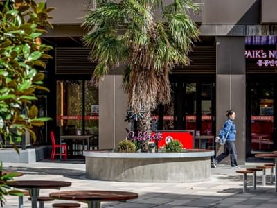 Woman walks past a Melbourne noodle restaurant with outdoor seating and a central palm tree.