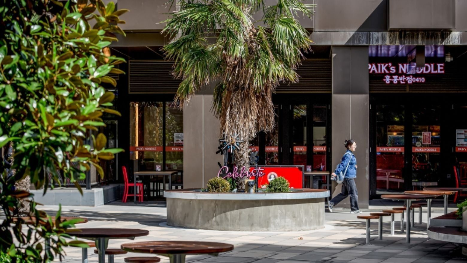 Woman walks past a Melbourne noodle restaurant with outdoor seating and a central palm tree.