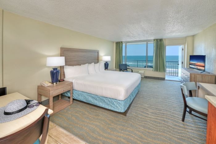 A wide-angle view of a hotel room with a bed, TV, balcony, and ocean view.