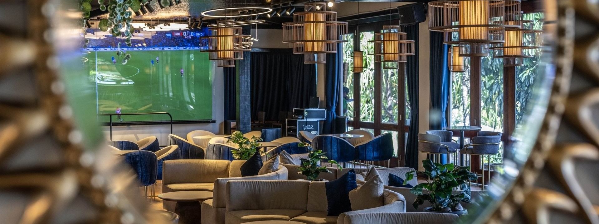 Grey sofas by round tables under cage pendant lights near a soccer screen in Hibiscus Lounge at Warwick Fiji Resort and Spa