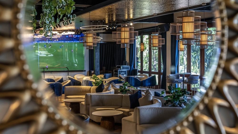 Grey sofas by round tables under cage pendant lights near a soccer screen in Hibiscus Lounge at Warwick Fiji Resort and Spa
