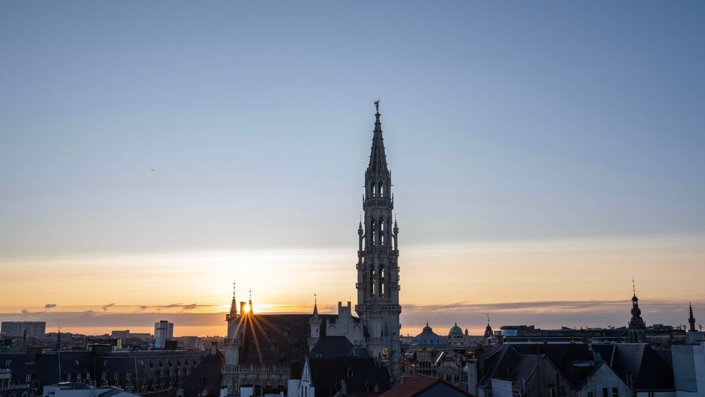 Sunset over Brussels cityscape with prominent spire and buildings silhouetted against orange sky.