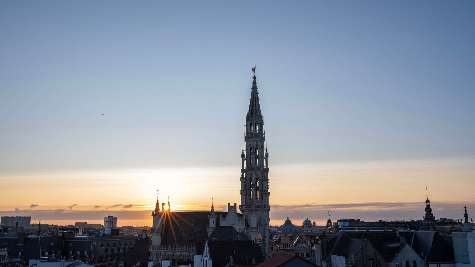 Brussels skyline at sunset with ornate Gothic tower rising above historic rooftops under a blue and golden sky.