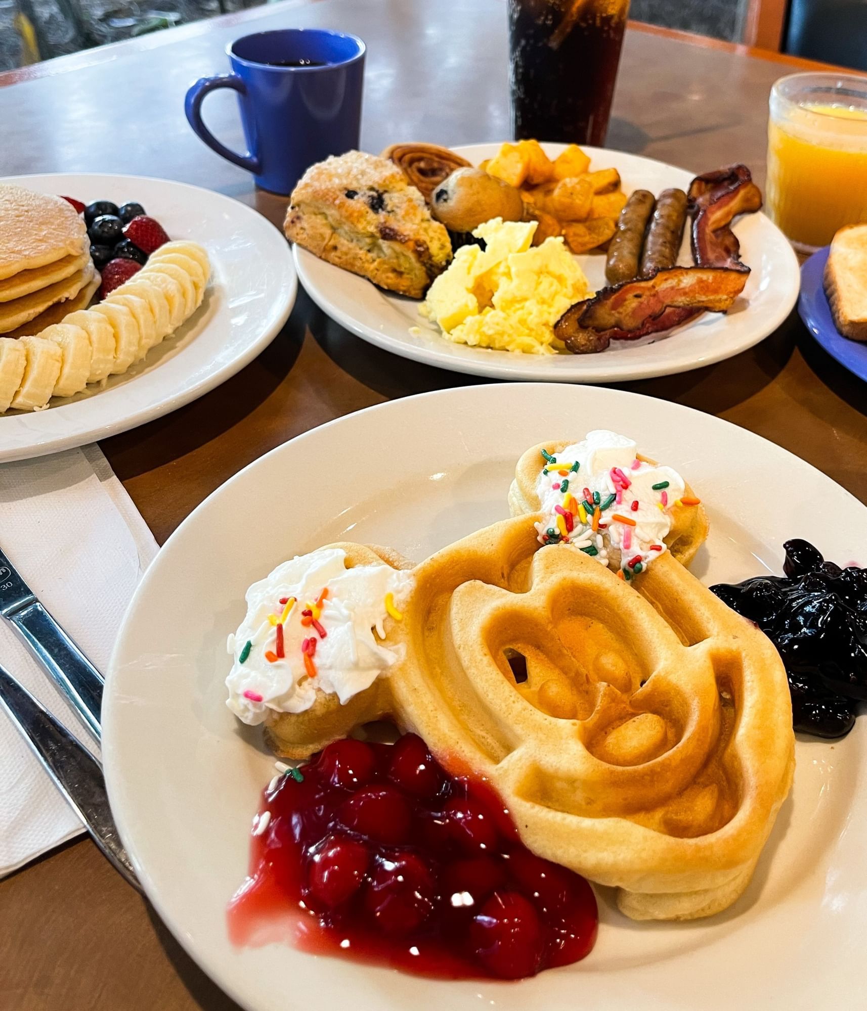 A plate with a Mickey Mouse-shaped waffle topped with whipped cream, sprinkles, and sides of cherry and blueberry compote sits among other breakfast items like pancakes, scrambled eggs, bacon, sausage, a scone, juice, and tea. 