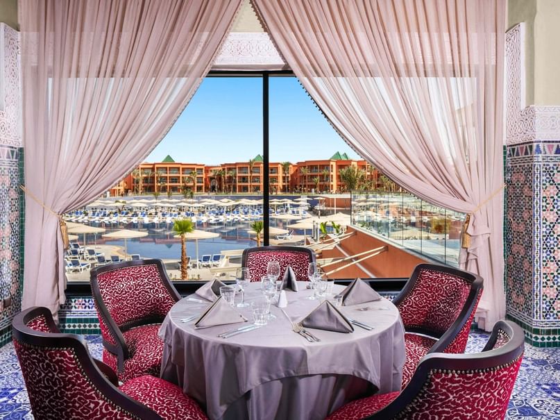 Elegantly set dining table with wine glasses and napkins surrounded by red chairs, overlooking a resort.