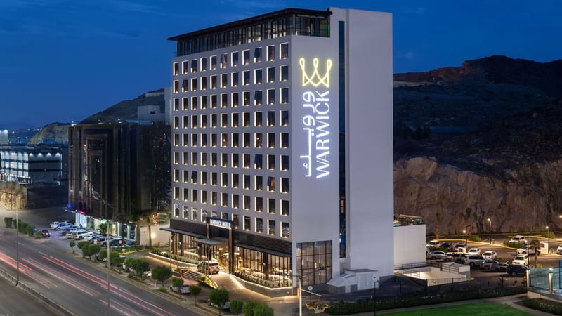 Exterior view of the Warwick Al Taif Hotel at night with illuminated signage and surrounding urban landscape