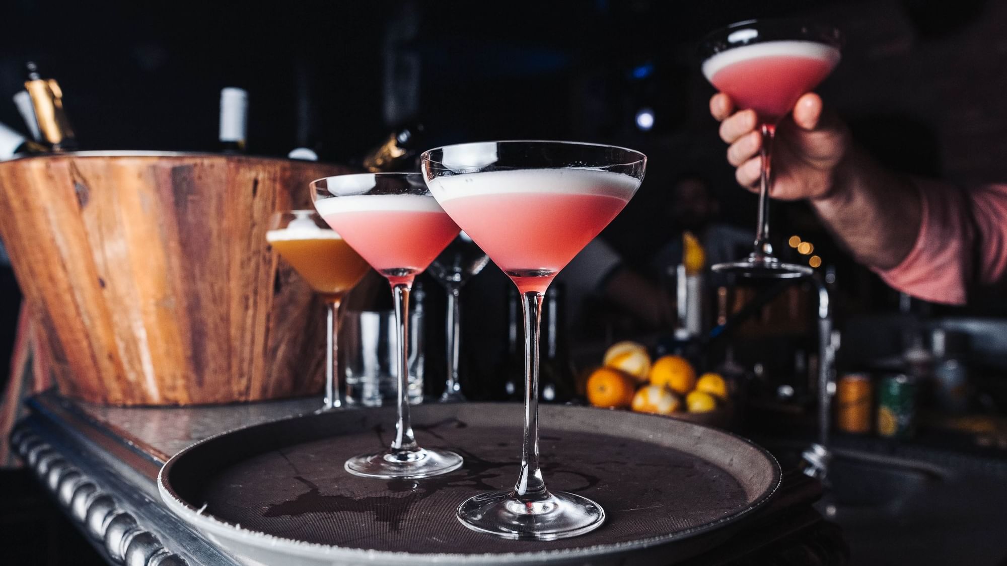 Three elegant pink cocktails on a round tray, as a person serves another drink from behind a wooden bar at Paramount Hotels
